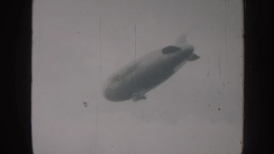 An airship with visible tail fins and gondola tracks upper-left across a hazy sky in 8mm home-movie footage consistent with late-1950s amateur cine practice.
