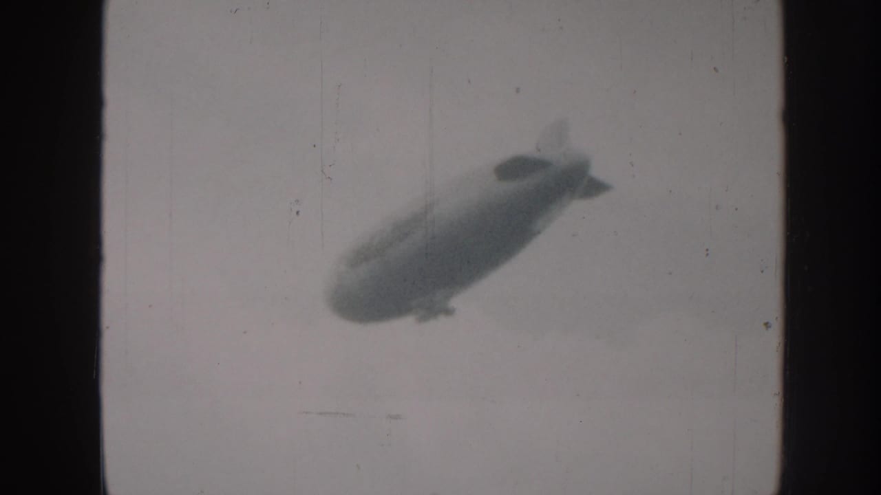 Monochrome airship shifted to upper-right of frame, continuing across overcast sky in grainy 8mm film.