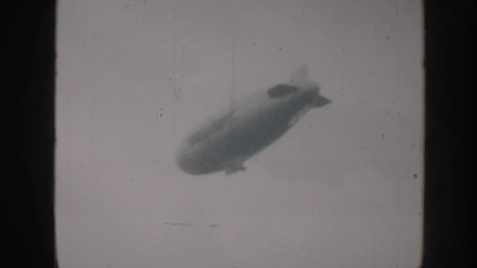 Blimp with tail fins and gondola continues upper-right across a hazy sky in 8mm home-movie footage; consistent with late-1950s amateur filmmaking in Nebraska.