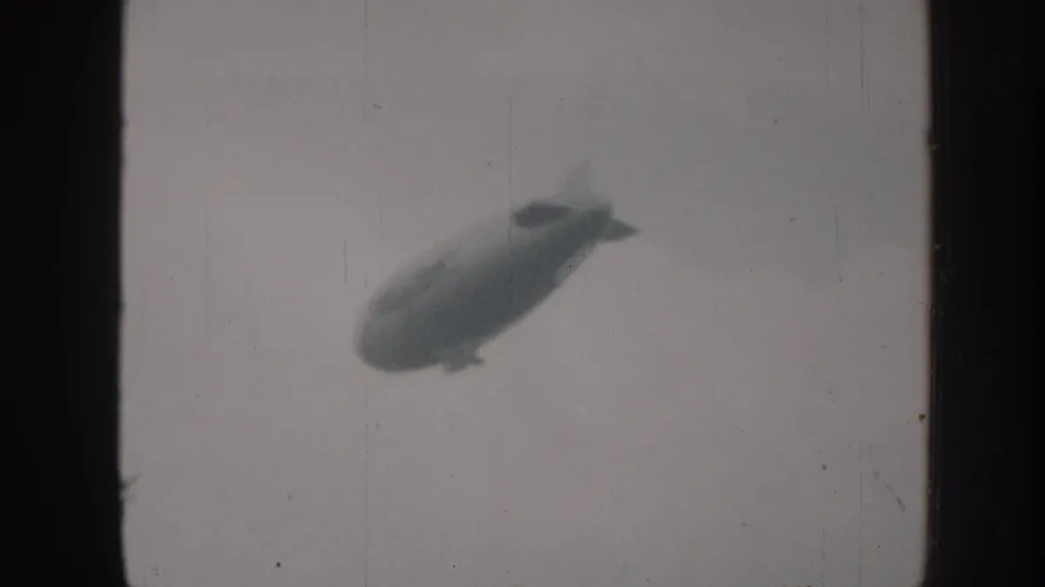 Airship with tail fins and gondola drifts through overcast sky in grainy monochrome 8mm home-movie footage; the sampled clip ends on this aerial view.