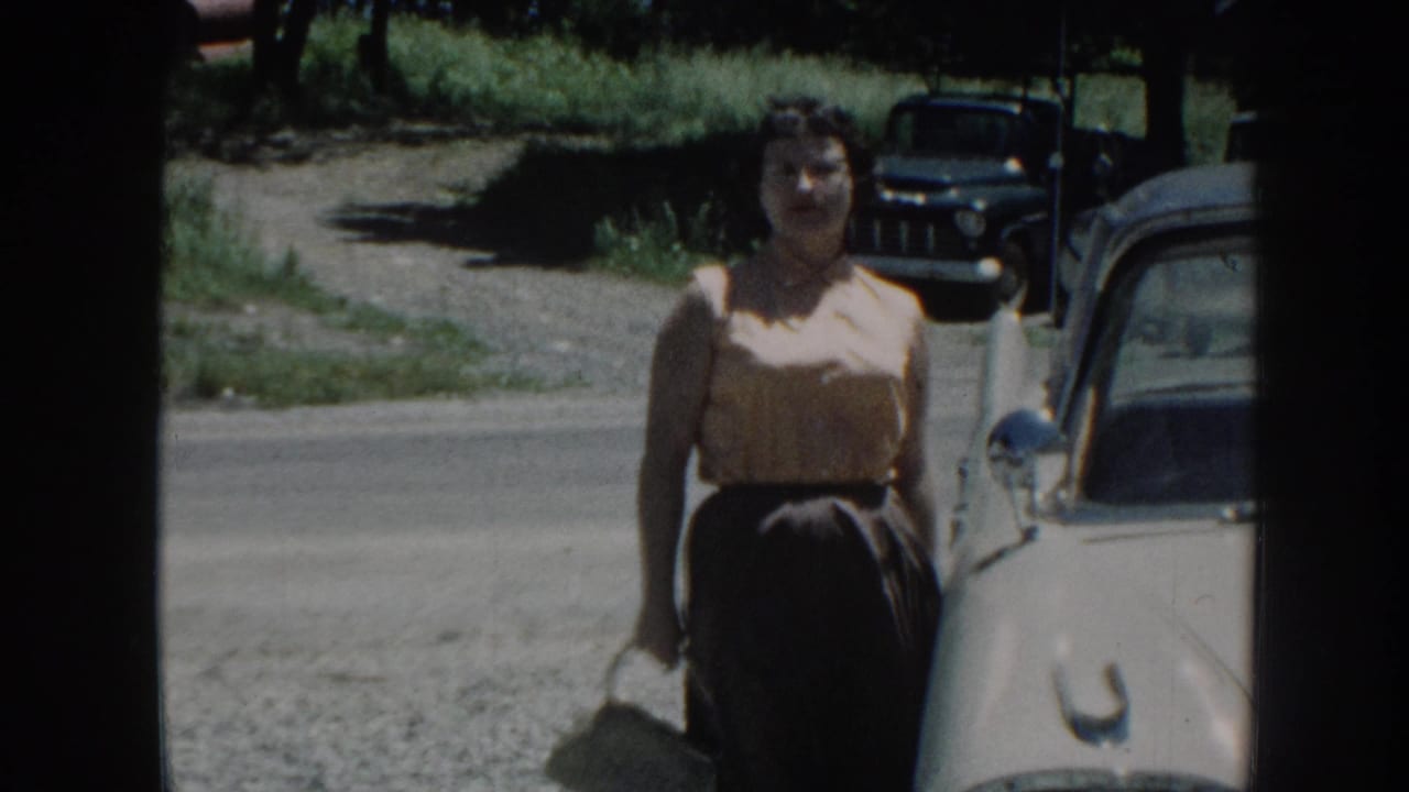 Woman in 1950s dress stands facing camera beside a cream-colored vintage American sedan; gravel road and green summer foliage visible, Nebraska, 1958.