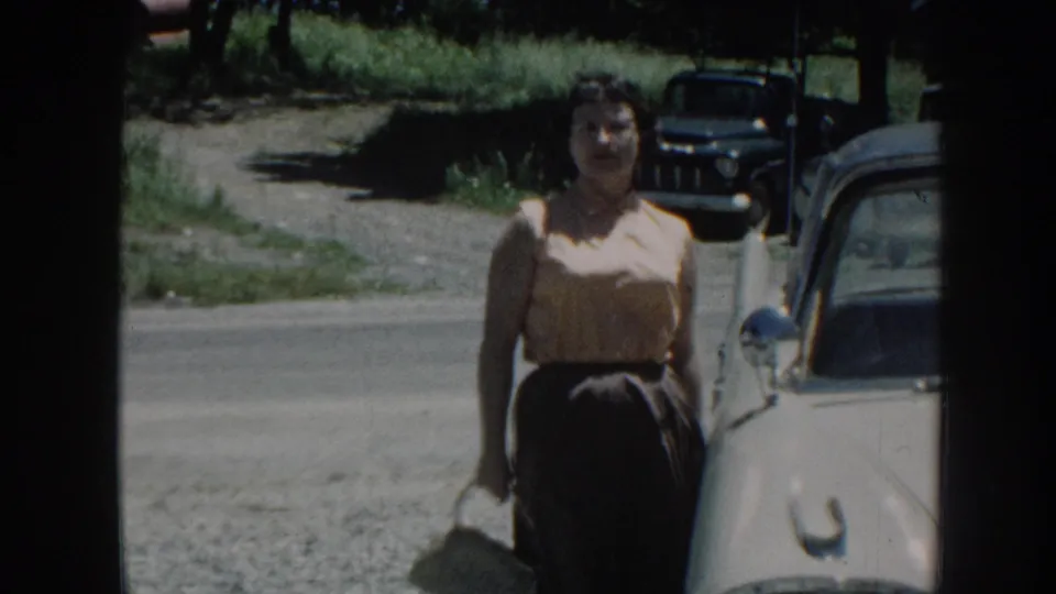 Woman in 1950s dress stands facing camera beside a cream-colored vintage American sedan; gravel road and green summer foliage visible, Nebraska, 1958.