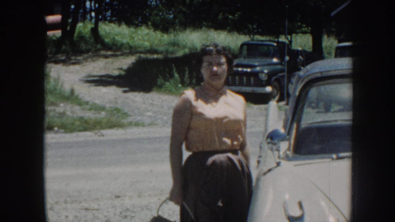Woman in yellow blouse and dark skirt facing camera; cream vintage car to her right and dark vintage truck visible in background on a gravel road.