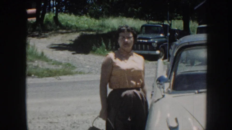 Woman in 1950s attire faces camera between a cream-colored vintage car and a dark vintage truck; gravel road and summer foliage visible, Nebraska, circa 1958.