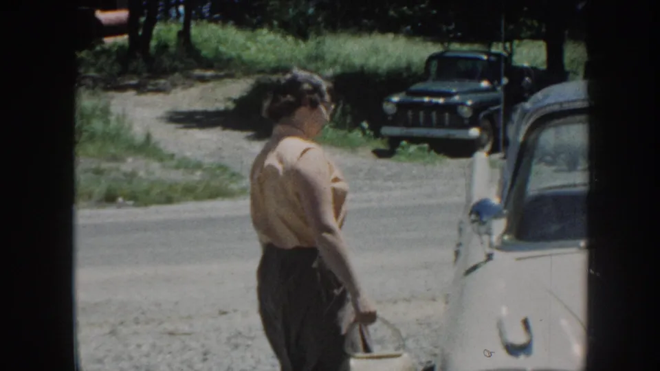 Woman in 1950s dress seen in profile carrying a small pail or handbag beside a cream vintage sedan; dark truck and gravel road visible, Nebraska, circa 1958.