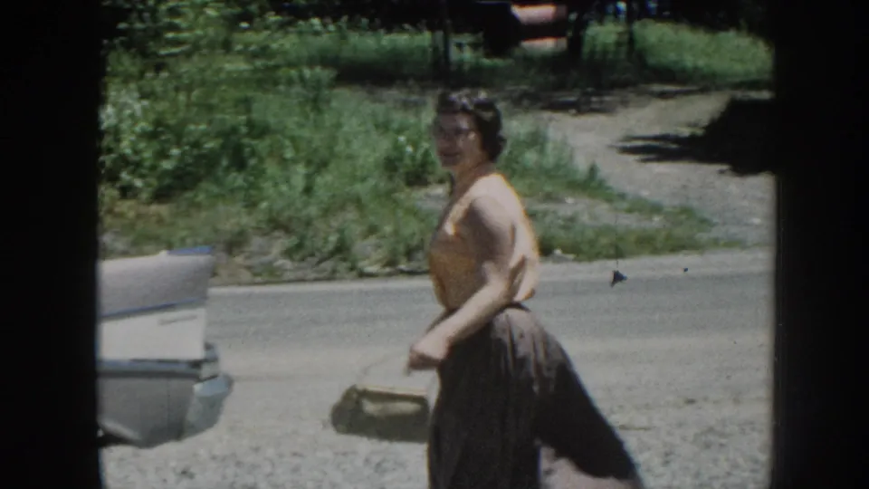 Woman in 1950s attire walks away while glancing back over her shoulder, carrying a small handbag beside a parked cream vintage sedan, Nebraska, circa 1958.