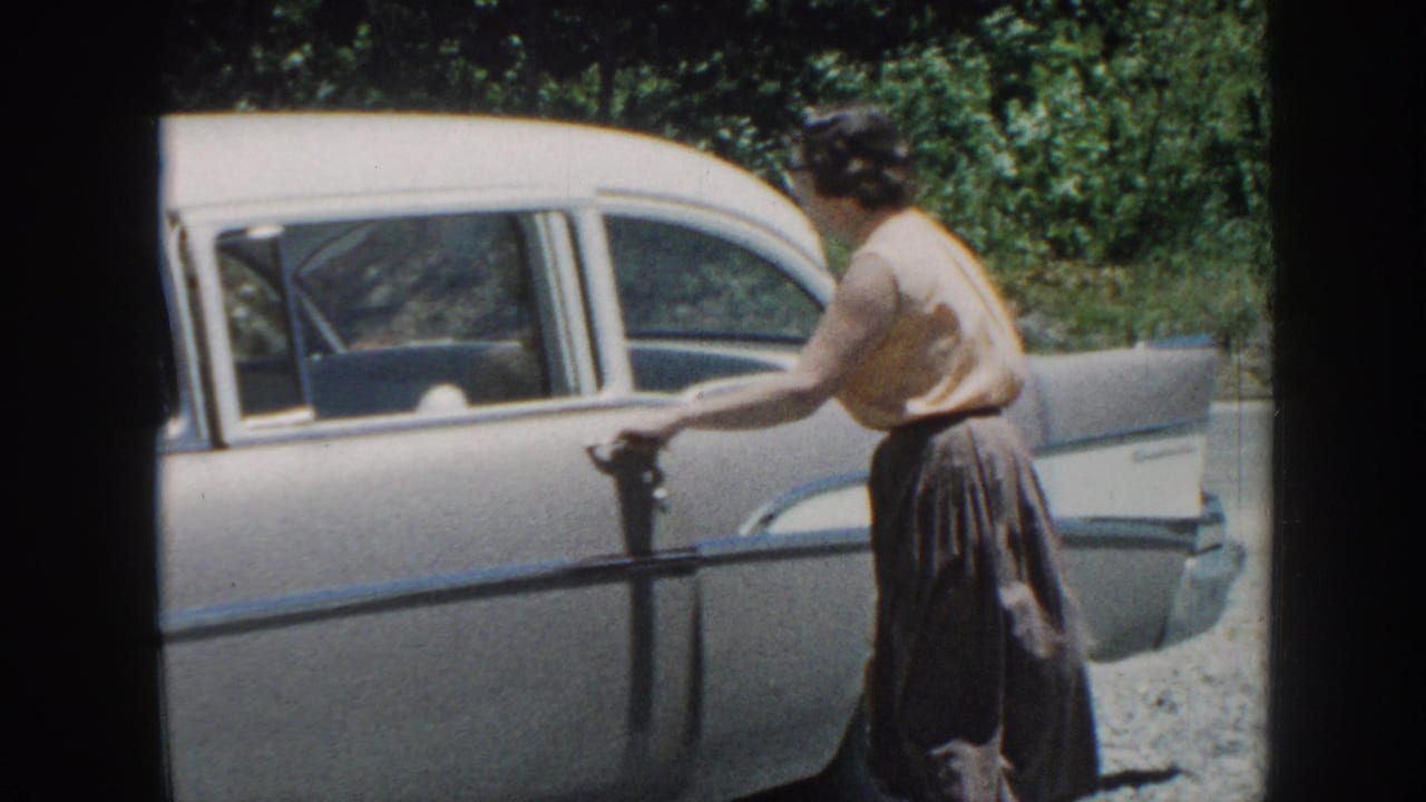 Woman in light top and dark skirt pulling open the rear door of a pale American sedan, green foliage in background.