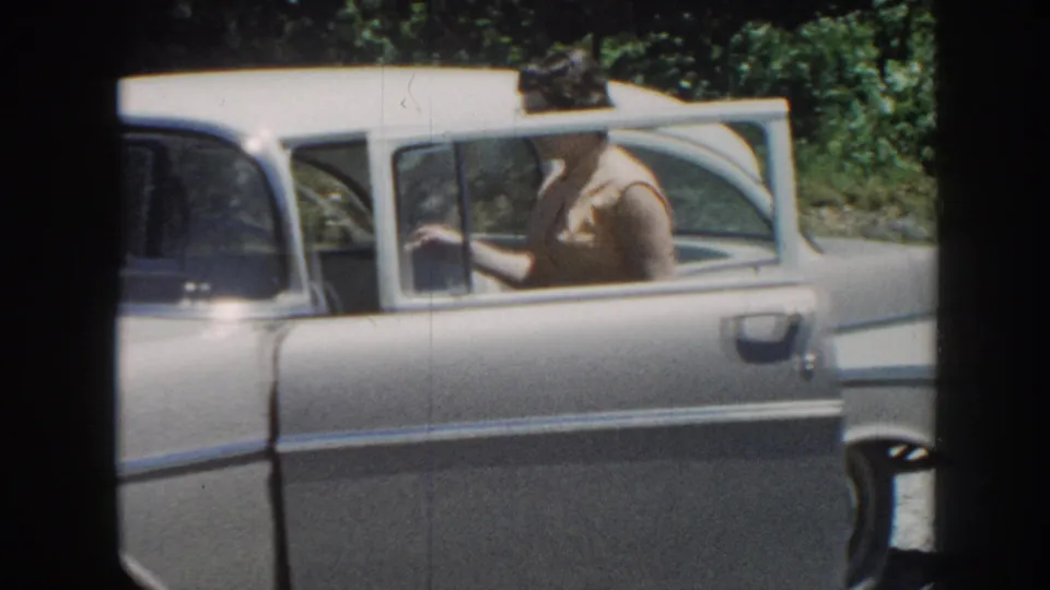 Woman entering the rear passenger seat of a late-1950s American sedan, circa 1958 8mm color home movie, Nebraska; a second figure visible in background.