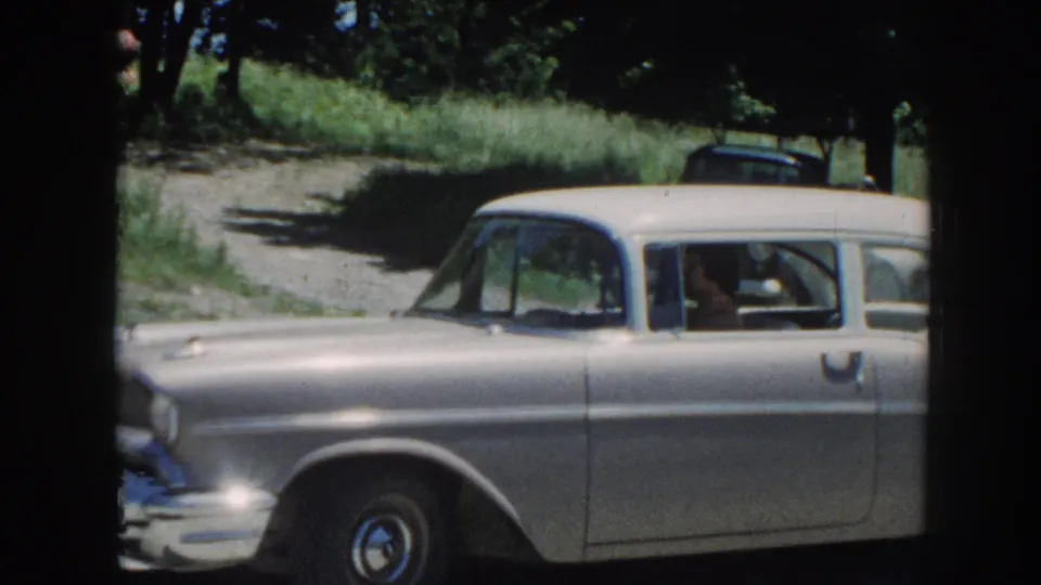 Large pale American sedan with prominent tailfins and whitewall tires on a gravel driveway, circa 1958 Nebraska 8mm home movie; occupant visible through window.