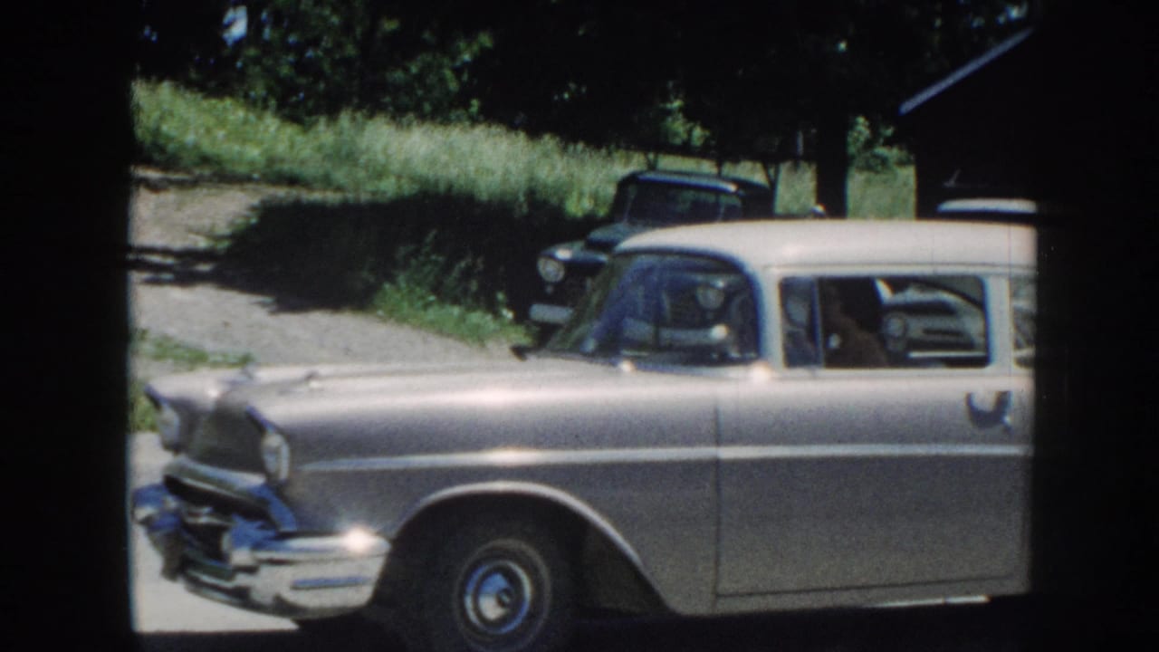 8mm home-movie frame showing a silver late-1950s American station wagon in a gravel yard in Nebraska, with a dark vintage pickup truck visible behind, circa 1958.