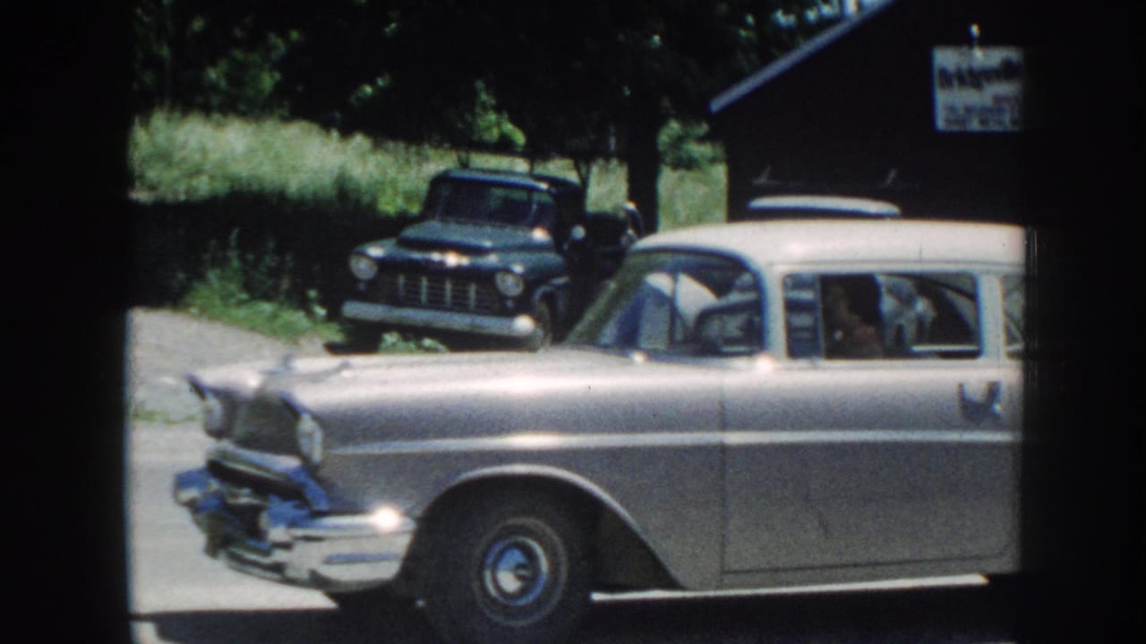 Silver late-1950s station wagon at front-three-quarter angle; dark pickup truck behind; partial commercial sign visible upper right; leafy trees.