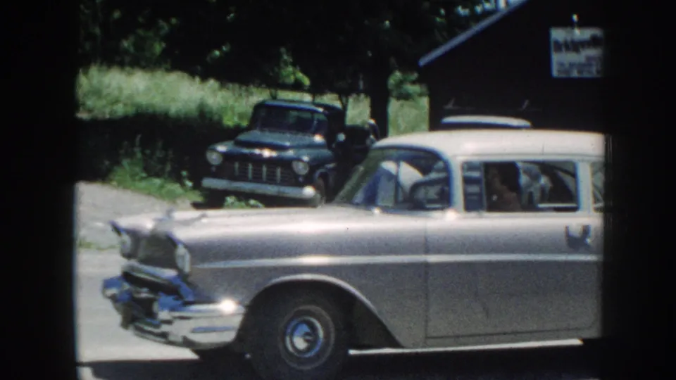 8mm home-movie frame of a silver late-1950s American station wagon with a dark vintage pickup and a partially legible commercial sign in the background, Nebraska, c. 1958.