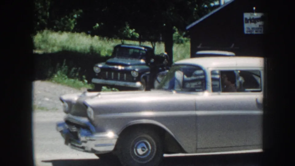 Home-movie frame showing both a silver late-1950s station wagon and a dark vintage pickup in a shared yard, with a partially legible sign on a structure behind, Nebraska, c. 1958.