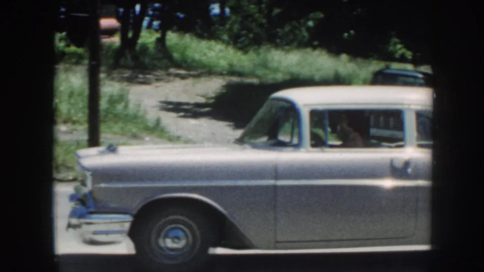 8mm home-movie frame showing the side profile of a silver late-1950s American station wagon with a person visible through the driver's window, Nebraska, c. 1958.