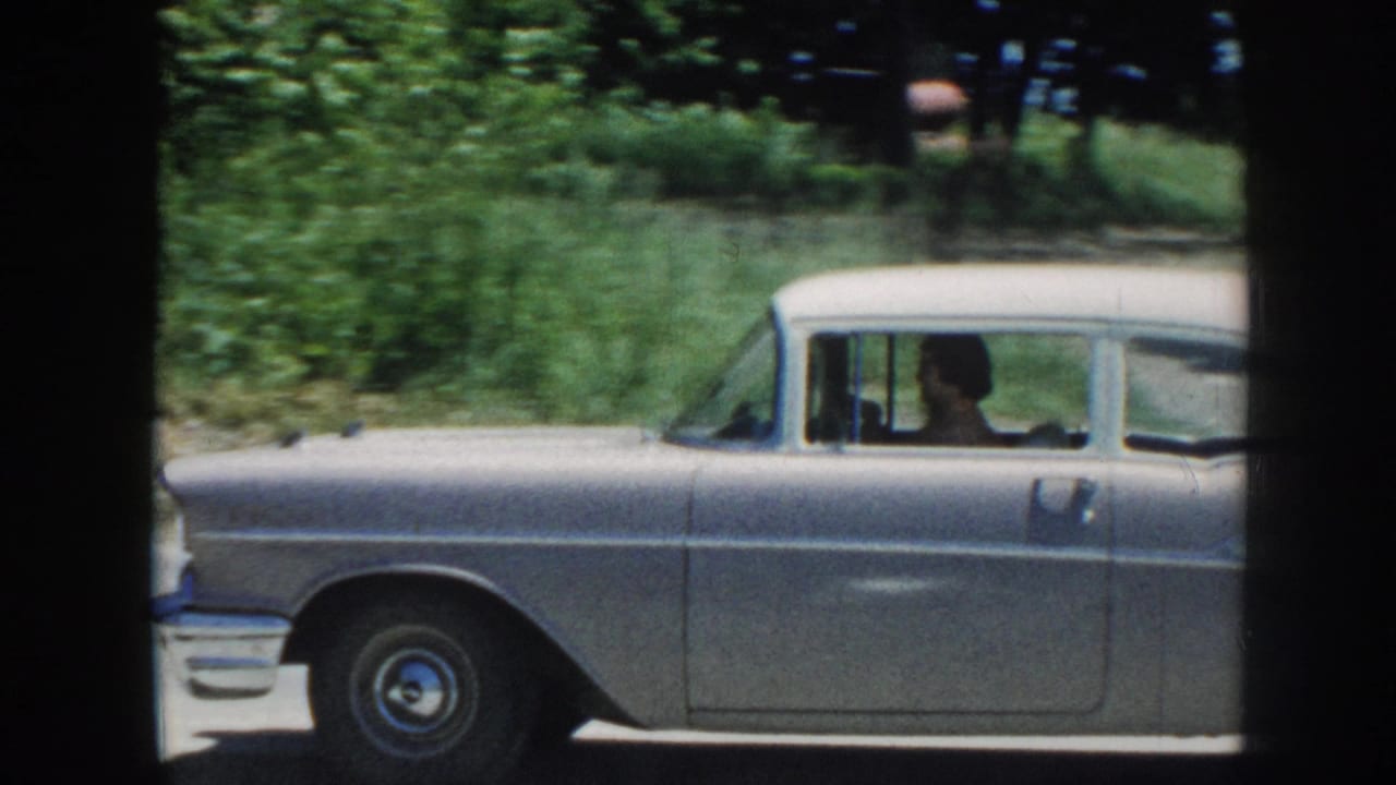 A silver late-1950s American sedan moves along a tree-lined road, driver visible through windshield; 8mm home movie, Nebraska, 1958.