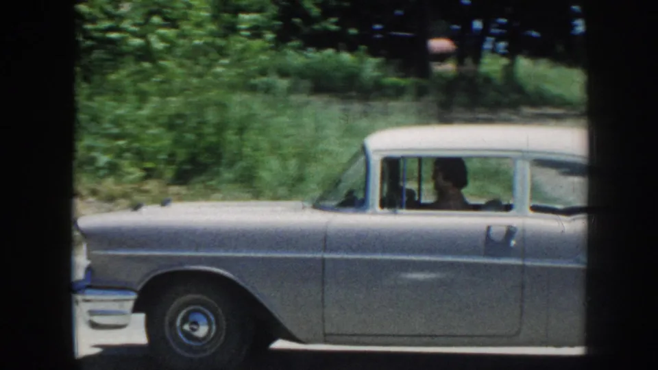 A silver late-1950s American sedan moves along a tree-lined road, driver visible through windshield; 8mm home movie, Nebraska, 1958.