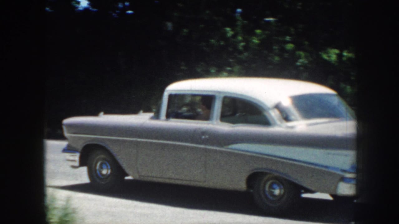 Rear view of a silver late-1950s American sedan passing along a tree-lined road; 8mm home movie, Nebraska, 1958.