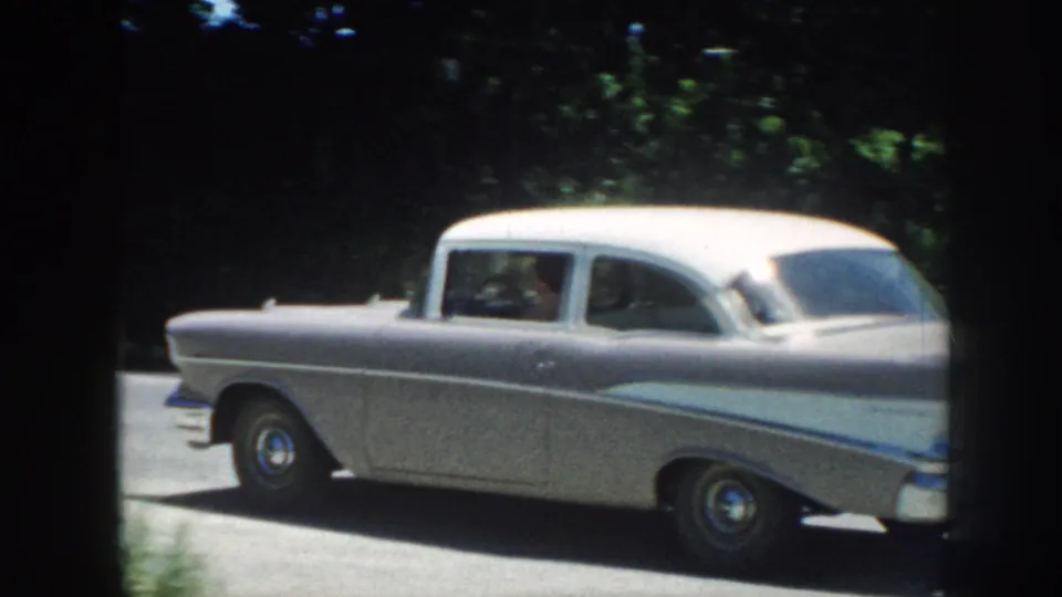Rear view of a silver late-1950s American sedan passing along a tree-lined road; 8mm home movie, Nebraska, 1958.