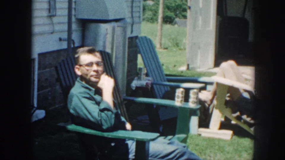 A middle-aged man in a green shirt and glasses relaxes in a green Adirondack chair in a sunlit backyard; 8mm home movie, Nebraska, 1958.