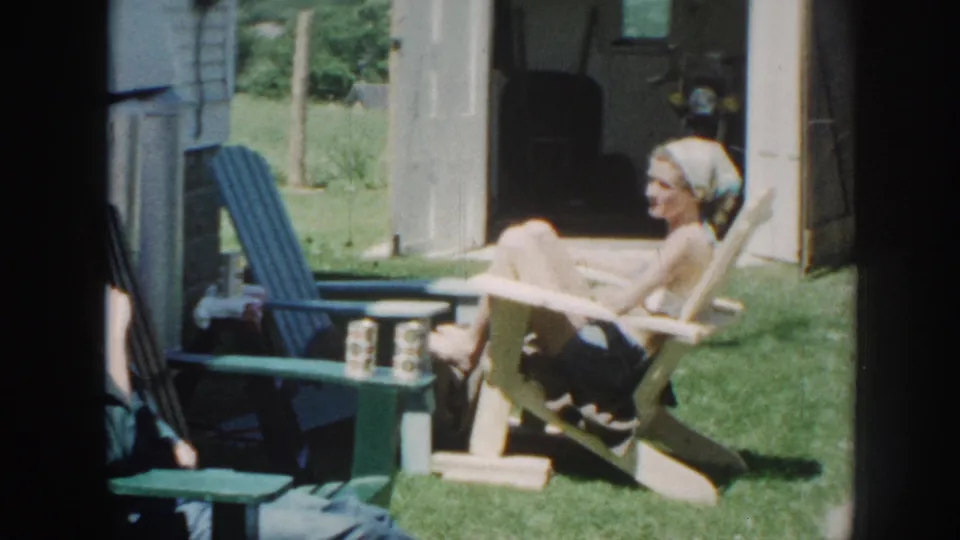 A woman wearing a headscarf relaxes in a wooden Adirondack chair in a sunlit backyard beside a house; 8mm home movie, Nebraska, 1958.