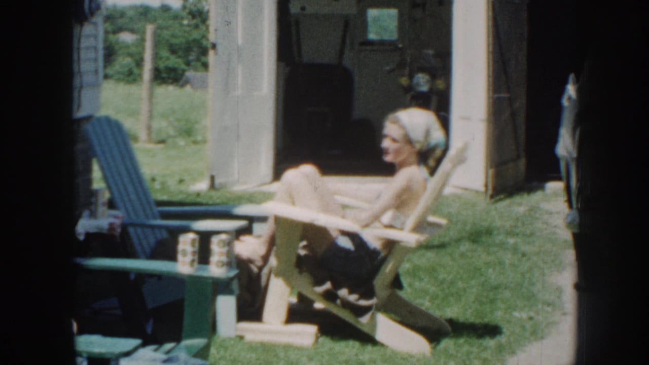 A woman in a light headscarf and dark shorts laughs in a wooden Adirondack chair in an apparent Nebraska backyard, circa 1958, 8mm home movie.