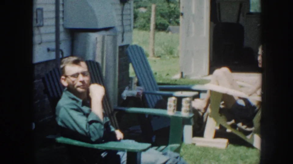 A man wearing horn-rimmed glasses and a green shirt sits in a blue Adirondack chair in a sunny residential backyard, Nebraska circa 1958, 8mm home movie.
