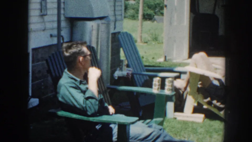 A man in horn-rimmed glasses and a green shirt relaxes in a blue Adirondack chair in a rural backyard, Nebraska circa 1958, 8mm home movie.