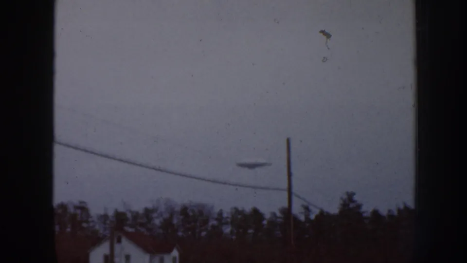 Overcast sky over a Nebraska rural landscape with utility poles, evergreen treeline, and white building with red roof; a small unidentified dark shape appears in the upper sky.