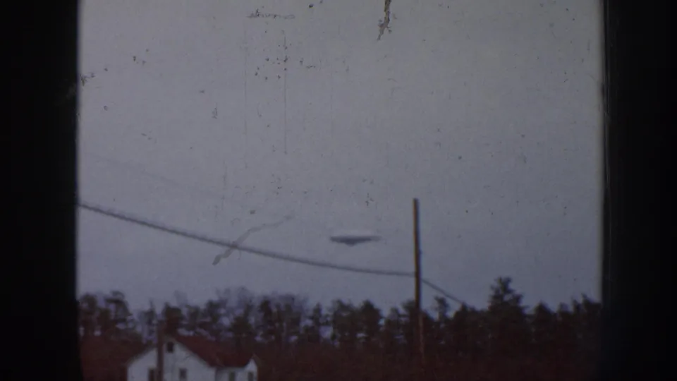 8mm footage of a cloudy sky over a rural landscape; emulsion scratches and dust spots are clearly evident, with a small dark shape again visible above the utility wires.