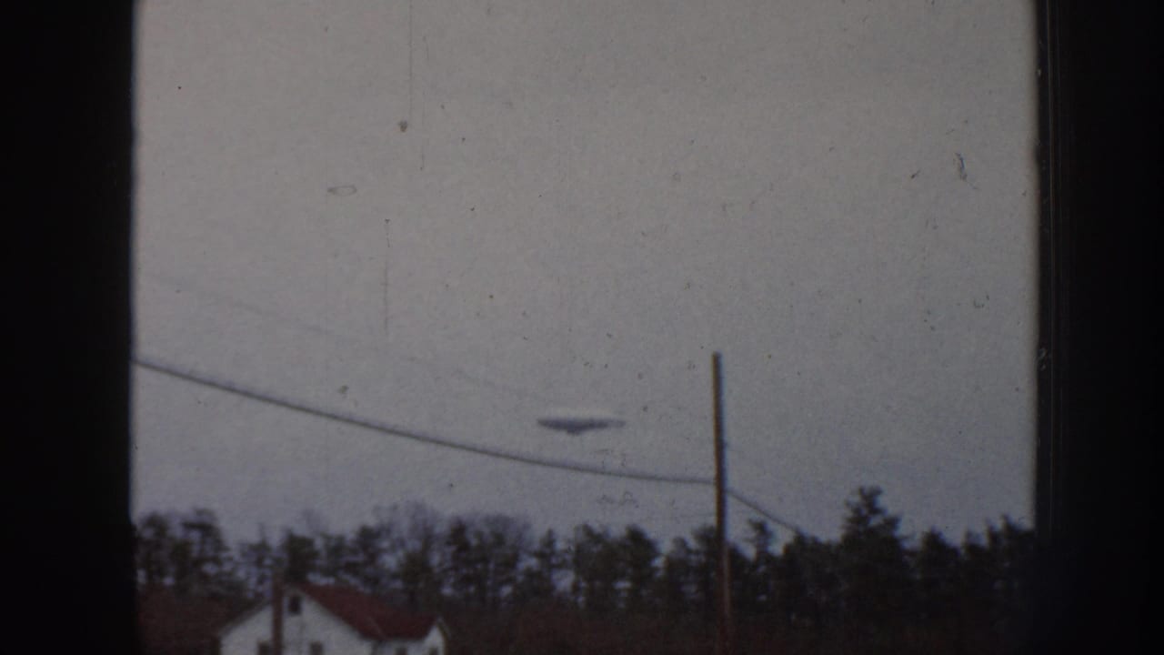 8mm home movie frame, Nebraska 1958, showing an unidentified disc-shaped aerial object in an overcast sky above a suburban treeline and partial red rooftop.
