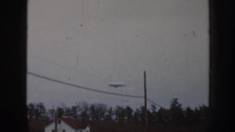 8mm home movie frame, Nebraska 1958, showing an unidentified disc-shaped aerial object in an overcast sky above a suburban treeline and partial red rooftop.