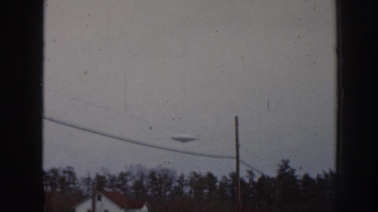 8mm film frame from 1958 Nebraska showing an unidentified aerial object in an overcast sky above a suburban treeline and red residential rooftop.