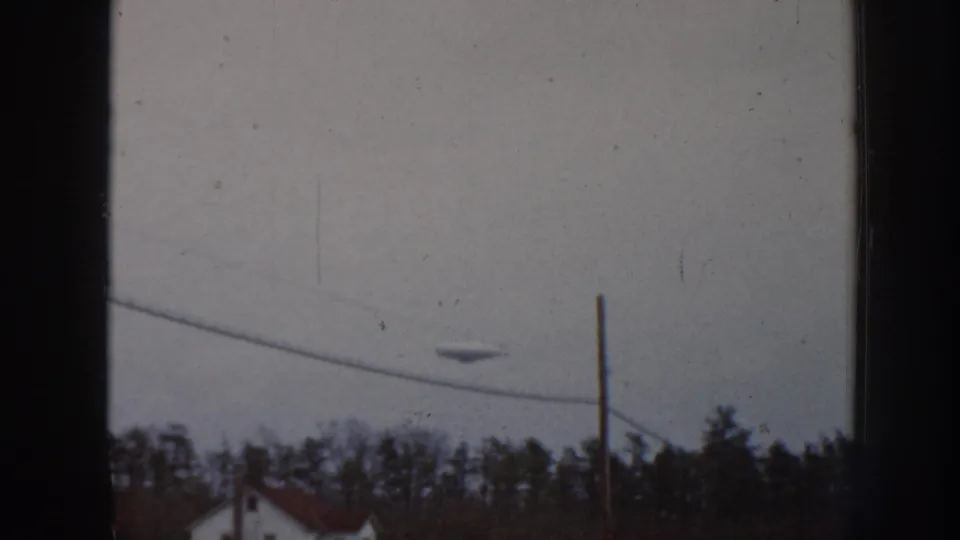 8mm film frame from 1958 Nebraska showing an unidentified aerial object in an overcast sky above a suburban treeline and red residential rooftop.