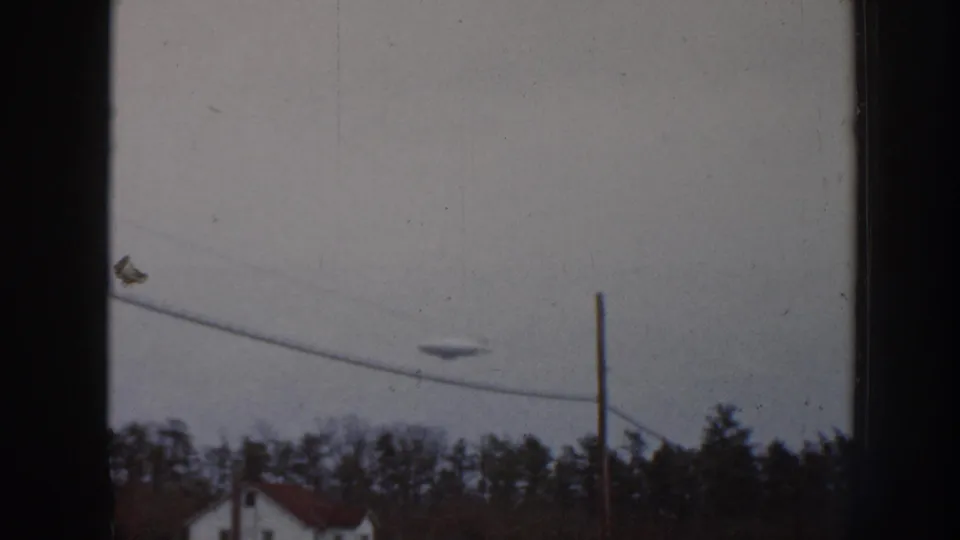 8mm home movie frame, Nebraska 1958; dark aerial object visible in overcast sky as camera angle shifts slightly downward, bringing the power line into closer view.