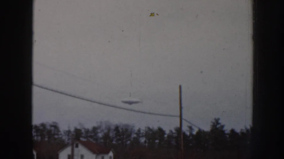 Final 8mm frame in the Nebraska 1958 sequence; unidentified aerial object above suburban roofline with a small film artifact visible in the upper right corner.
