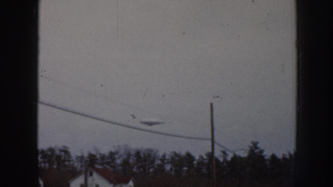 8mm home movie frame, 1958 Nebraska: small unidentified airborne object in overcast sky above diagonal power lines, conifers, and a single-story house.