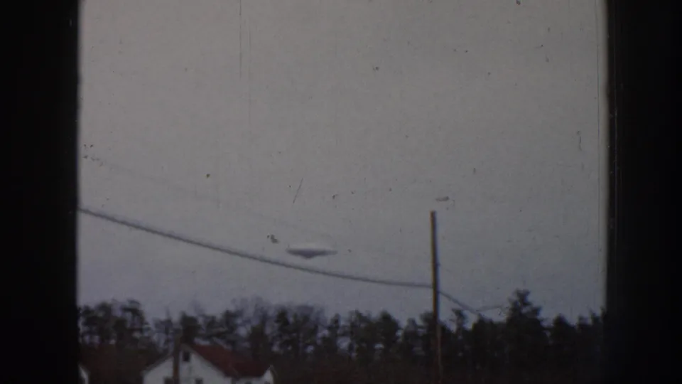 8mm home movie frame, 1958 Nebraska: small unidentified airborne object in overcast sky above diagonal power lines, conifers, and a single-story house.