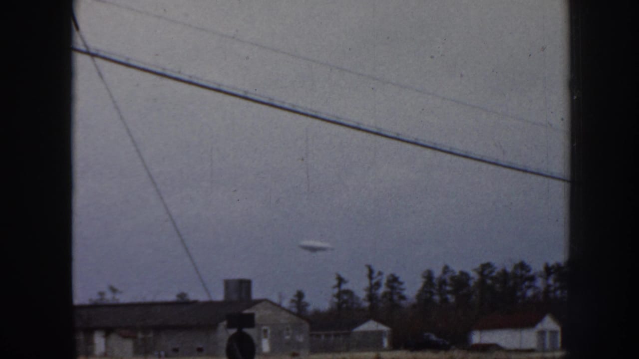 8mm home movie frame, 1958 Nebraska: pale airborne object near the horizon above mid-century residential structures and conifers under an overcast sky.