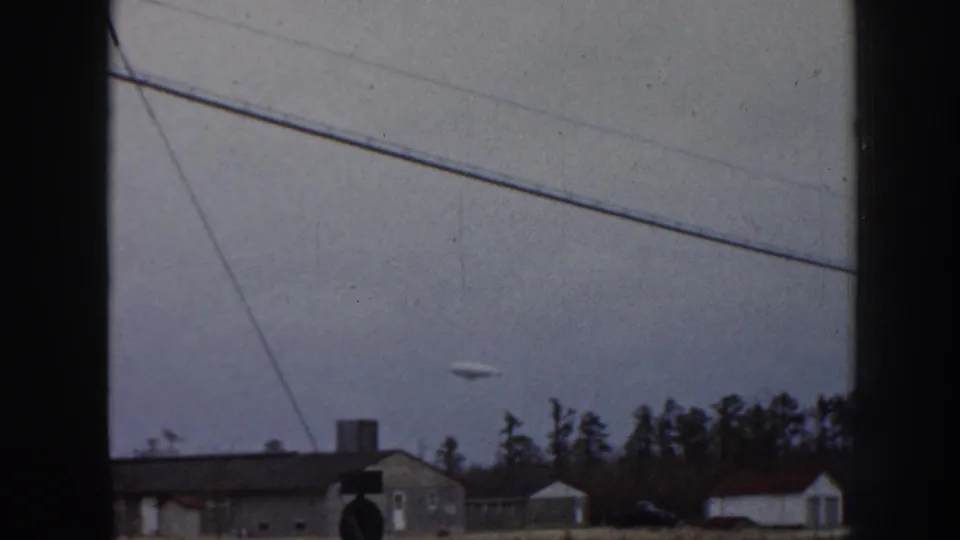 8mm home movie frame, 1958 Nebraska: pale airborne object near the horizon above mid-century residential structures and conifers under an overcast sky.