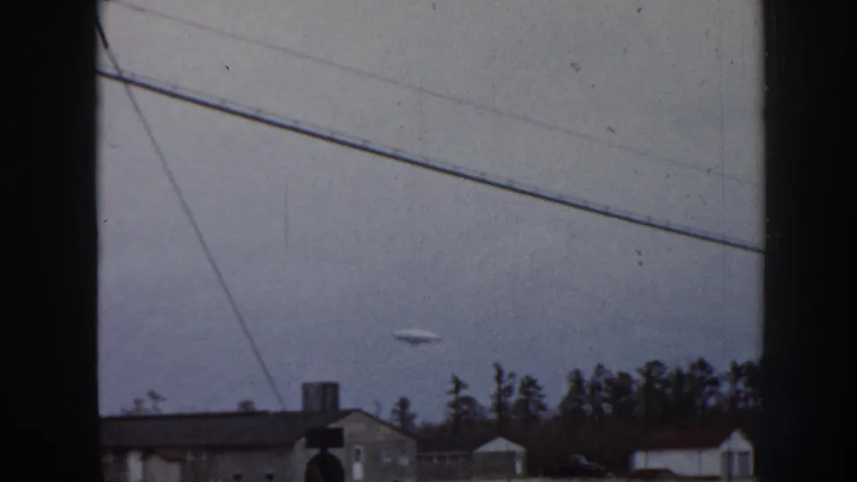 8mm home movie frame, circa 1958 Nebraska: flat pale aerial object above residential rooftops with chimneys, framed by diagonal power lines and a conifer treeline.