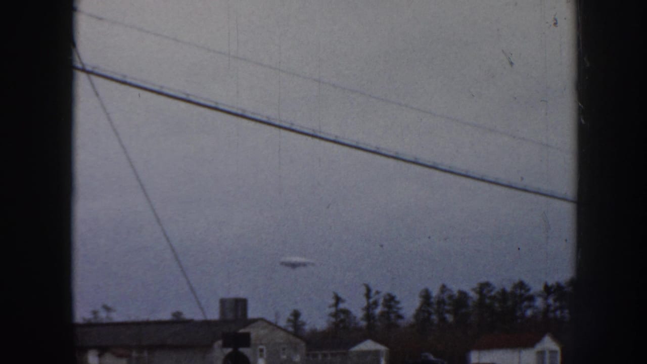1958 Nebraska 8mm home movie frame: overcast sky, diagonal power lines, and a small unidentified disc-shaped object visible above mid-century suburban houses and a conifer tree line.