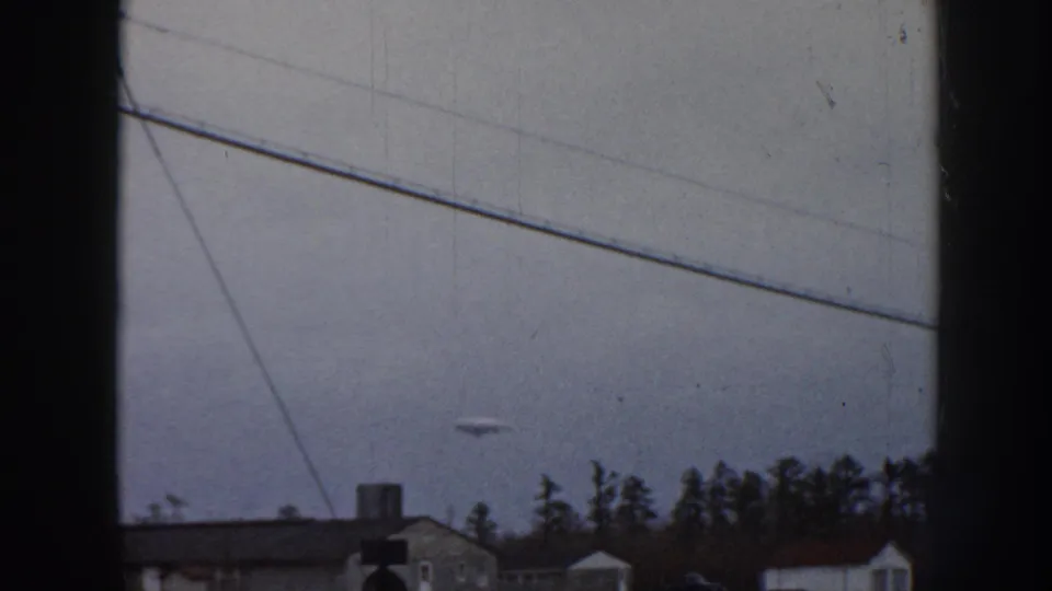 1958 Nebraska 8mm home movie frame: overcast sky, diagonal power lines, and a small unidentified disc-shaped object visible above mid-century suburban houses and a conifer tree line.