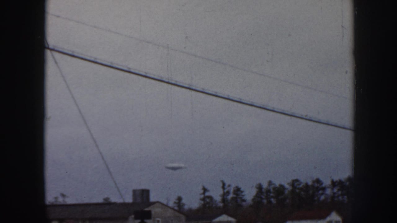 Second 1958 Nebraska 8mm sky frame showing the same persistent disc-shaped aerial object above mid-century suburban rooflines and conifers, consistent color fade and grain.
