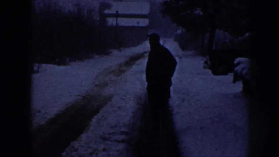 Dark, heavily blue-shifted 8mm frame from Nebraska, 1958: lone adult male silhouette on a snow-covered road, bare shrubs flanking both sides, fence or structure in background.