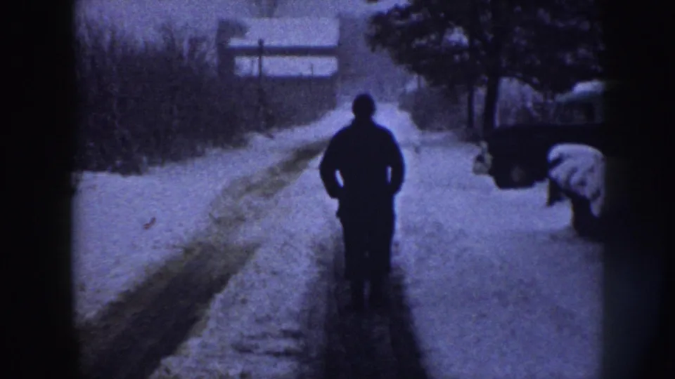 Late 1950s 8mm home movie frame from Nebraska: adult male walking away down a snow-covered path flanked by bare shrubs, with a snow-covered object at far right and a residential fence beyond.