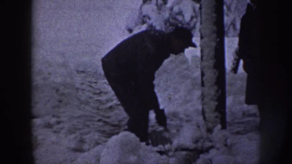 The man continues clearing snow from a sidewalk near a tree, with a partial figure at the right edge of the frame.