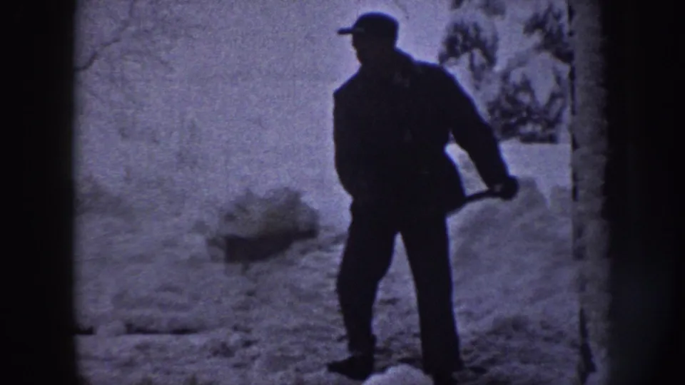 A man in a dark coat and cap stands with a shovel beside a snowy sidewalk in Nebraska, 1958.