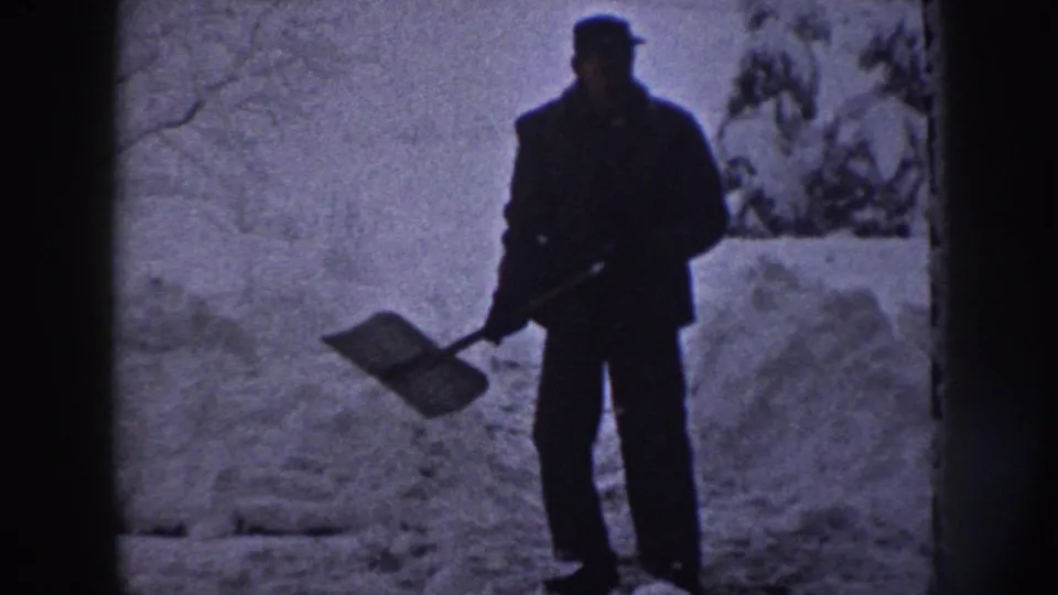In 1958 Nebraska home-movie footage, a man stands in deep snow with a shovel near trees and a residential path.