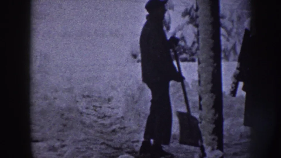 A man in winter clothing stands with a shovel in a snowy Nebraska residential yard, seen in soft 8mm silhouette.