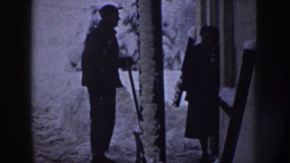 The man with the shovel stands near a woman in a long coat by a snow-covered residential exterior in Nebraska.